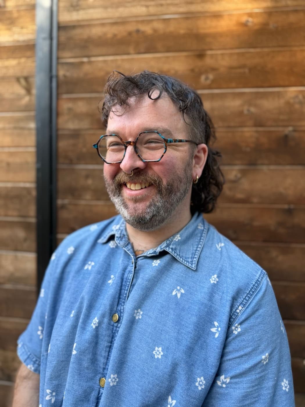 Jacob Perry, smiling, in a blue floral denim shirt against a cedar fence